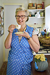 Anette Løkken står i sit retrobondehuskøkken med kaffekop og blå prikket kjole.