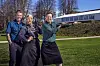 Tre værter i forklæder står i solen foran et hvidt bagedyst-telt i parken.