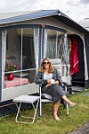 Kvinde sidder med kaffekop foran fortelt ved campingvogn på grøn plads.