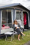 Kvinde sidder med kaffekop foran fortelt ved campingvogn på grøn plads.