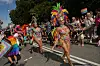 Strax-deltagere i fjerprydede kostumer danser under en pride-parade omgivet af regnbueflag.