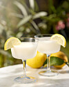 To lyse citrondrinks i coupeglas med citronskiver på et solrigt bord udendørs.
