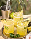 Tre gule cocktails med jalapeño og ananas på metalbakke i solrigt udemiljø.