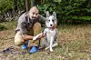 Deltager fra 'Alene i vildmarken' sæson 10 knæler i haven med sin hund på græsset foran træer.