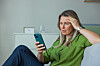 Kvinde sidder i sofa med hånden mod panden og ser bekymret på sin telefon.