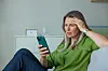 Kvinde sidder i sofa med hånden mod panden og ser bekymret på sin telefon.