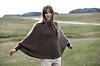 Kvinde i brun strikponcho står på en grøn mark med vej i baggrunden