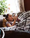 Person ligger på sofa under tæppe og kigger på smartphone i lys stue