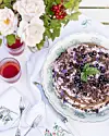 Lagkage med bær og chokolade på fad omgivet af blomster, glas og bestik på hvid dug.