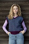 Person står foran trævæg iført mønstret strikket vest over langærmet bluse og jeans.