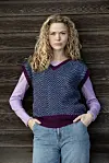 Person står foran trævæg iført mønstret strikket vest over langærmet bluse og jeans.