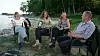 Fire personer sidder i stole og taler sammen ved en kyst med skov bagved.