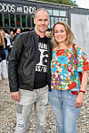 Par poserer smilende udendørs foran en bygning under en sommerfest.