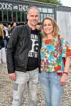 Par poserer smilende udendørs foran en bygning under en sommerfest.