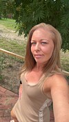 Kvinde tager selfie udendørs på terrasse foran have og træer.