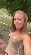 Kvinde tager selfie udendørs på terrasse foran have og træer.