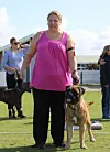 Kvinde i pink top står på græs med stor hund i snor ved hundearrangement.