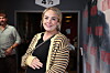 Smilende kvinde i stribet cardigan står ved Strax-fotovæg indendørs.