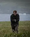 Mand i mørkt tøj går alene gennem en eng ved havet på en overskyet dag