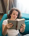 Kvinde ligger på turkis sofa og læser en bog i et lyst rum