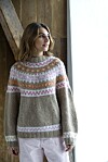 Kvinde står indendørs ved et vindue iført en beige sweater med farverigt rundt bærestykke.