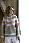 Kvinde står indendørs ved et vindue iført en beige sweater med farverigt rundt bærestykke.