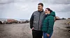 Par i jakker står arm i arm på en overskyet strand med havet bag dem.
