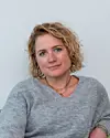 Kvinde med krøllet hår i grå trøje sidder foran en lys væg