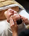 Behandler giver ansigtsmassage til person, der ligger på briks med hvidt håndklæde.