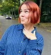 Person med rødt hår i blåstribet skjorte står på træomkranset vej.