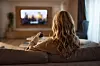 Kvinde set bagfra i sofaen, der holder en fjernbetjening og kigger på tændt tv i stuen.