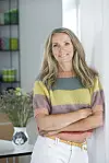 Kirstine Virén står i et lyst rum med farverig bluse og blomster i baggrunden.