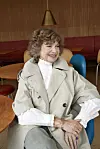 Ældre kvinde i beige frakke sidder smilende i farverig café