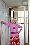 Ældre kvinde i lyserød trøje og blomstret nederdel står i en lys indendørs gang.