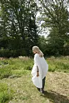 Kvinde i hvid kappe går på en eng foran en tæt grøn skov.