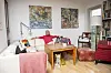 Ældre person sidder i hvid sofa i lys stue med malerier og bogreol
