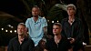 Fire mænd sidder udendørs om aftenen foran palmer og lyskæder i et tv-setup.