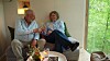 Ældre par skåler med vin i sofa ved stort vindue med udsigt til skov.