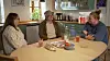 Tre personer taler sammen ved et køkkenbord med drikkevarer og snacks