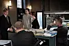Efterforskere og advokat taler sammen ved et rodet skrivebord på et mørkt kontor.
