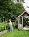 Kvinde plukker blomster i højbede ved åbent drivhus i grøn have