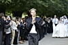Model i sort jakkesæt går på udendørs catwalk med publikum langs træomkranset allé.