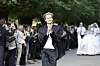 Model i sort jakkesæt går på udendørs catwalk med publikum langs træomkranset allé.