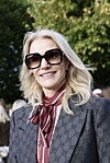 Kvinde i Gucci-jakke og store solbriller smiler ved et udendørs arrangement.