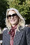 Kvinde i Gucci-jakke og store solbriller smiler ved et udendørs arrangement.