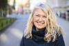 Smilende kvinde med lyst bølget hår står på en rolig bygade i blødt lys.