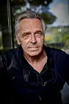Nærbillede af Michael Falch ved pressemøde foran glasfacade med grøn baggrund.