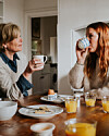 To kvinder drikker fra krus ved et morgenbord med juice og tallerkener i et lyst køkken.