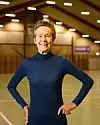 Ældre kvinde i mørk trøje står i en sportshal med hænderne i siden.