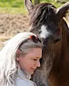 Kvinde med gråt hår står tæt og rører panden mod en brun hest udendørs.